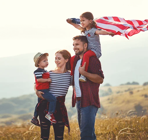 A joyful family poses outdoors, with a child holding a flag, against a scenic landscape of rolling hills under a bright sky.