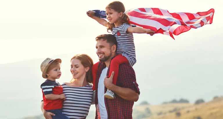 A joyful family poses outdoors, with a child holding a flag, against a scenic landscape of rolling hills under a bright sky.