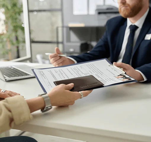 A person hands a business card holder to another during a meeting, with a clipboard and documents visible on the table.