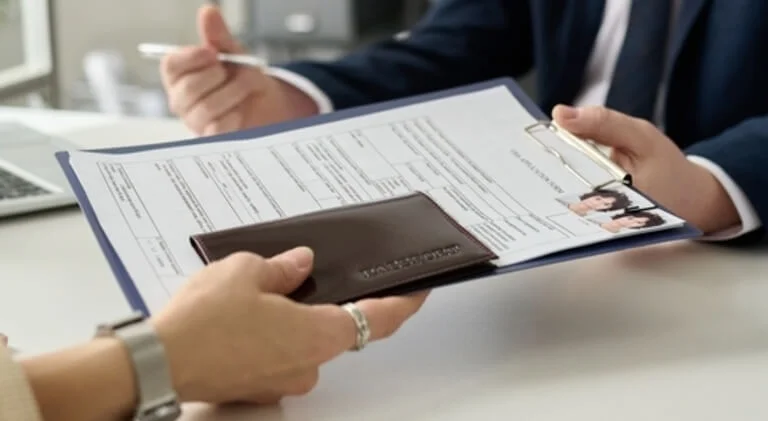 A person hands a business card holder to another during a meeting, with a clipboard and documents visible on the table.