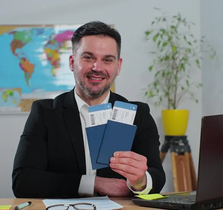 Smiling man in a suit sitting at a desk, holding up two boarding passes with passport covers, with a world map and plant in the background.