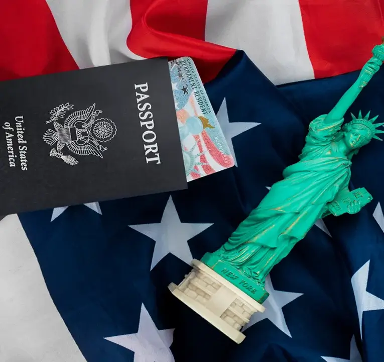 U.S. passport with a green card partially visible, placed on an American flag next to a small Statue of Liberty figurine labeled 'New York'.