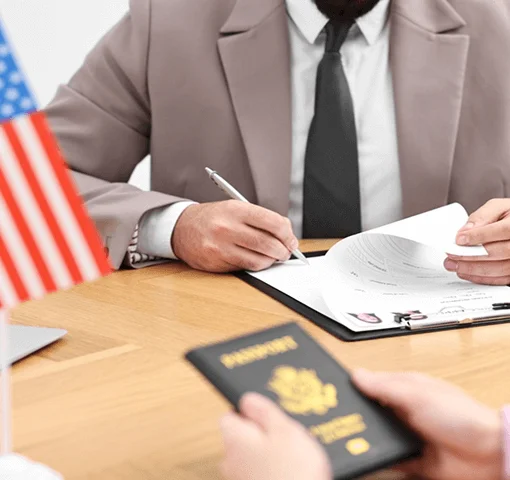 A man's hands hold documents while he signs paperwork on a clipboard, with a small American flag and a passport visible on the table.